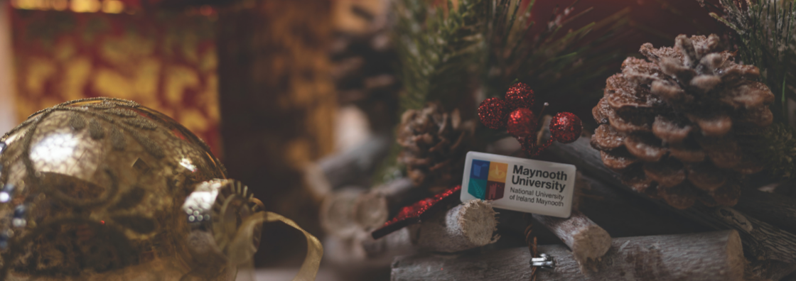 Maynooth University logo enamel pin against a background of Christmas lights and decoration