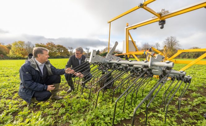 2 men squatting down in a field, behind a yellow seed robotic planter