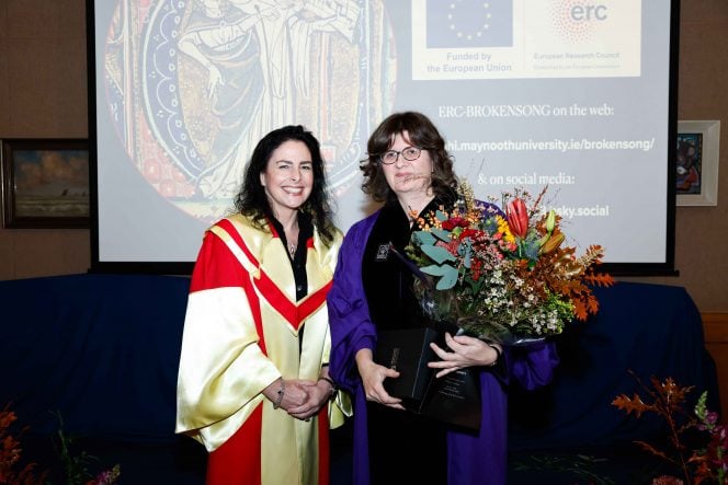 Woman in yellow robes stands next to a woman in a purple gown holding a large bunch of flowers