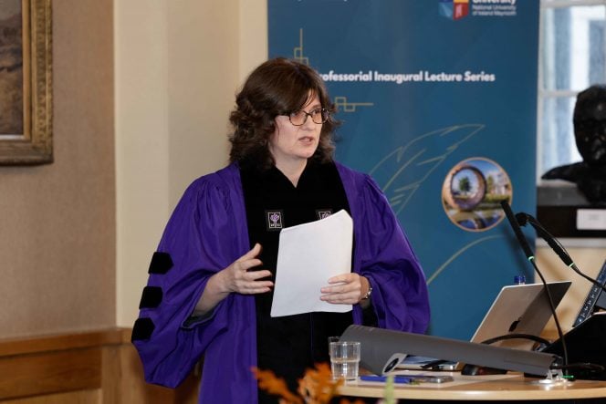 Woman with black hair wearing a purple gown standing in front of a blue banner with a sheet of paper in her hand