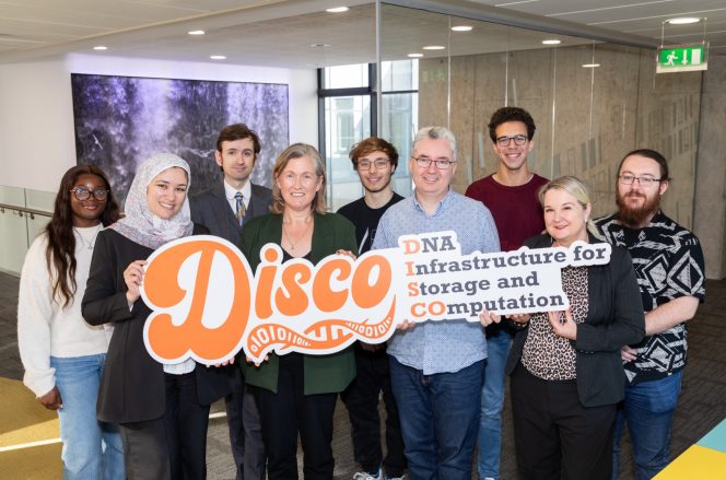 4 women and 4 men posing in front of foamex advertising board with the word DISCO in orange font. 