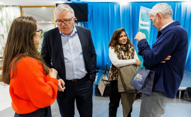 2 women and 2 men, women on the left with brown hair and red jacket; man in navy suit and light blue shirt, second women on the middle right in a white shirt, leopard print scarf, black pants and man on the right in navy blue cardigan and grey pants