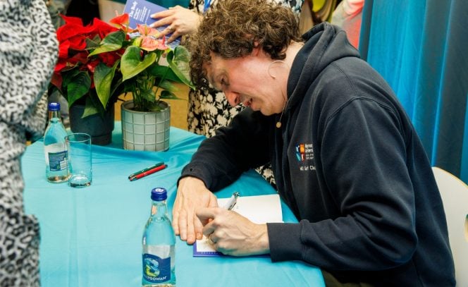 Man in navy hoodie signing a book. Light blue table cloth, and red poinsettia flower in background