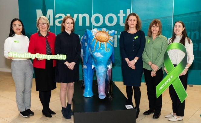 6 women in the picture, left women in white top, and grey pants, women in a red jacket and black skirt, women 3 in all black outfit, pictured with a colourful Elephant mental health sculpture depicting Darkness into Light, with another woman in all black outfit, next woman in a green blouse and dark pants and woman on far right in white top and dark pants. Also holding signs depicting 'Health Campus 