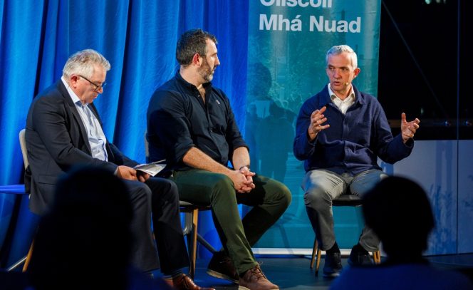 3 men on a panel discussion, blue background, left man in a navy suit, white shirt, middle man wearing navy shirt and green trousers and man to the right wearing a navy cardigan 