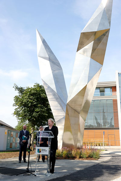 Dr Alison FitzGerald stands at a clear plexiglass podium in front of the stainless steel ‘Freedom’ sculpture by Alexandra Wejchert