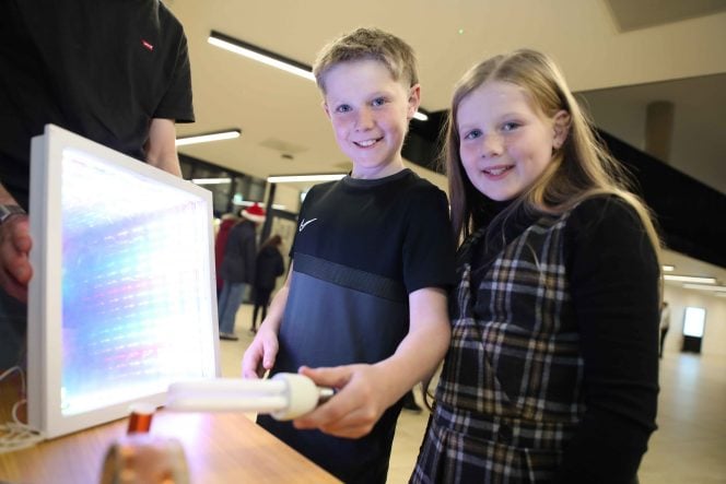 Boy in black t-shirt and girl in tartan pinafore stand in front of a screen