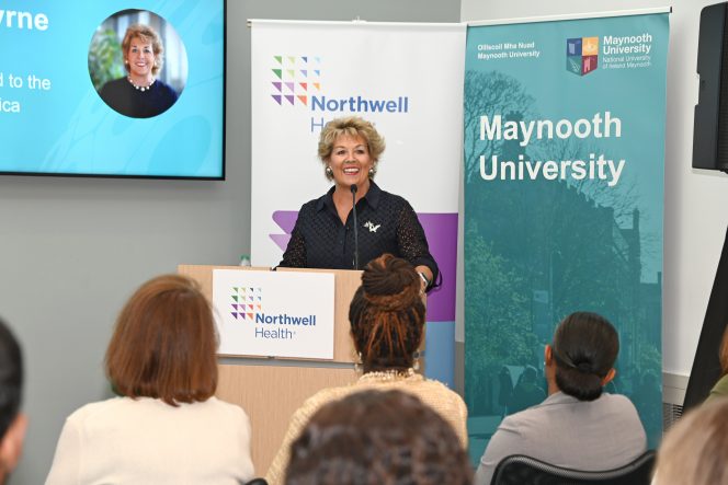 Woman with blond curly hair at a lectern with two banners behind her for Northwell Health and Maynooth University