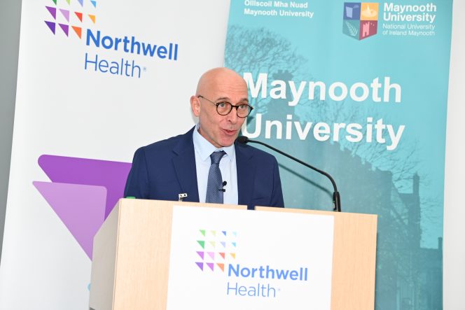 Man in a blue suit at a lectern in front of two banners reading Northwell Health and Maynooth University