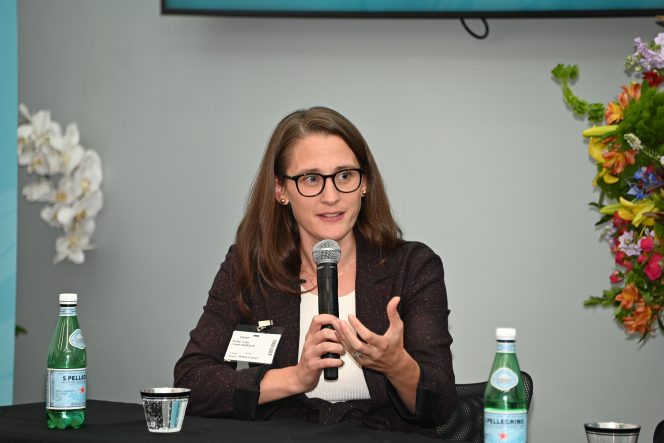 Women in a brown jacket with long hair and glasses holding a microphone