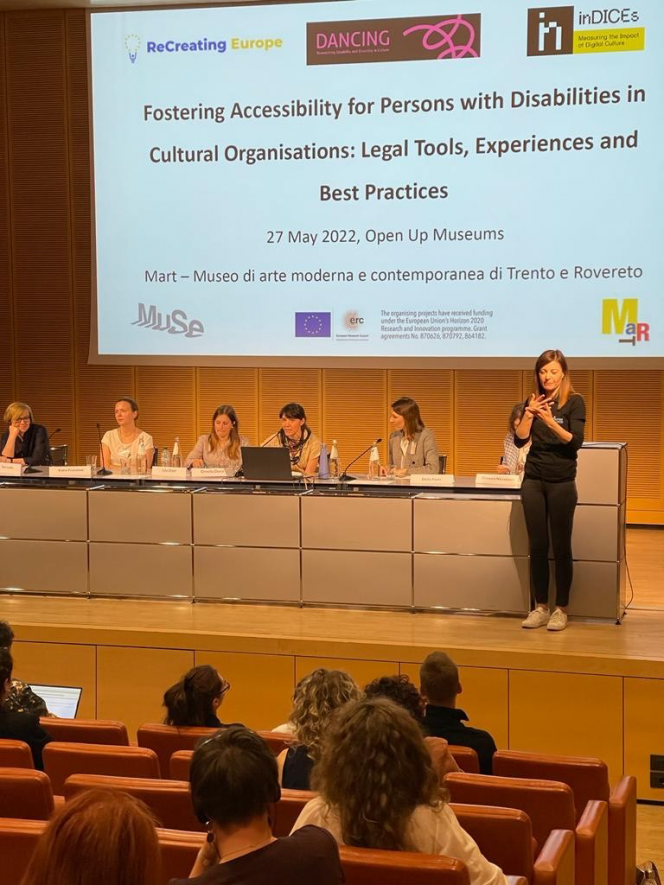 6 women sitting behind a table presenting at a conference with a sign language interpreter in front. Screen in the background shows 3 project logos: ReCreating Europe, DANCING and inDICEs title Fostering Accessibility for Persons with Disabilities in Cultural Organisations: Legal Tools, Experiences and Best Practices. 27 May 2022, Open Up Museums. Funding body logos on the bottom - EU flag, ERC logo 