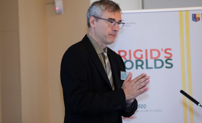 Man in a black jacket with hands joined standing before a banner reading Brigid's Worlds