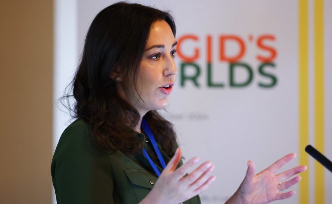 Woman with dark hair and green dress speaking before a banner reading Brigid's Worlds