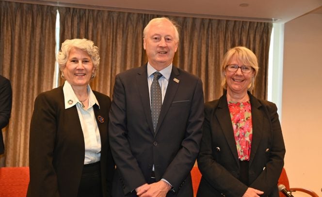 Woman with blond curly hair, man in a black suit and woman with a red blouse standing in a row