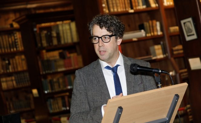 Man with curly hair and glasses in a jacket, shirt and tie standing in front of a lectern with bookshelves in the background