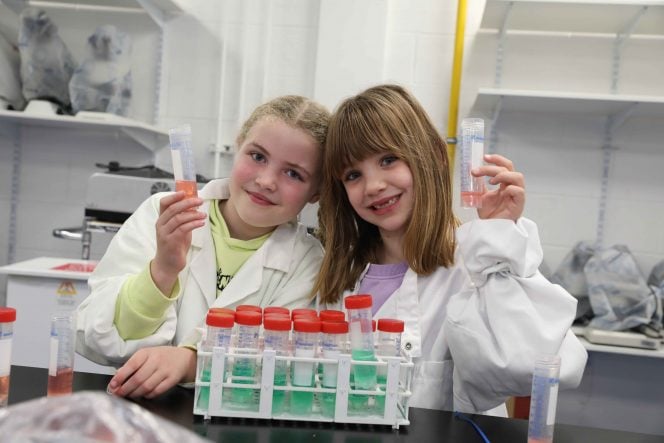 Girl with fair hair and girl with sandy hair wearing white coats hold test tubes