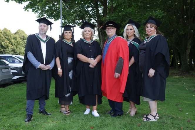 Group of six graduates standing on grass in front of trees in robes and mortar board hats