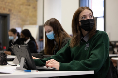 Four female students in school uniform wearing covid masks in a bright room. Two of the students in background in front of exposed red brick wall using laptops Two of the girls are in the foreground one is looking at their laptop the other student in the very front is looking up.