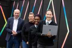 From left to right A man, a woman and two female secondary school students in uniform smiling at the camera and standing in front of a black background with a colorful linear design. The student on the right is holding a laptop.
