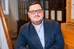 man with glasses and beard wearing a navy suit jacket and baby blue shirt posing against wooden stair rail