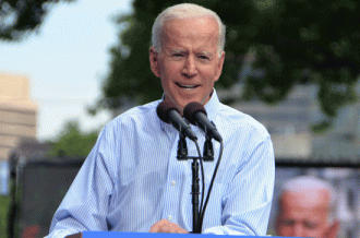 Joe Biden, visible from the waist up, in a blue shirt stands at a podium and speaks into the microphone, he is smiling