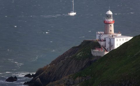 White and red lighthouse building sitting on a rocky outcrop with a boat approaching by sea