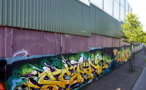 Wall with grafitti on the bottom topped by a high fence