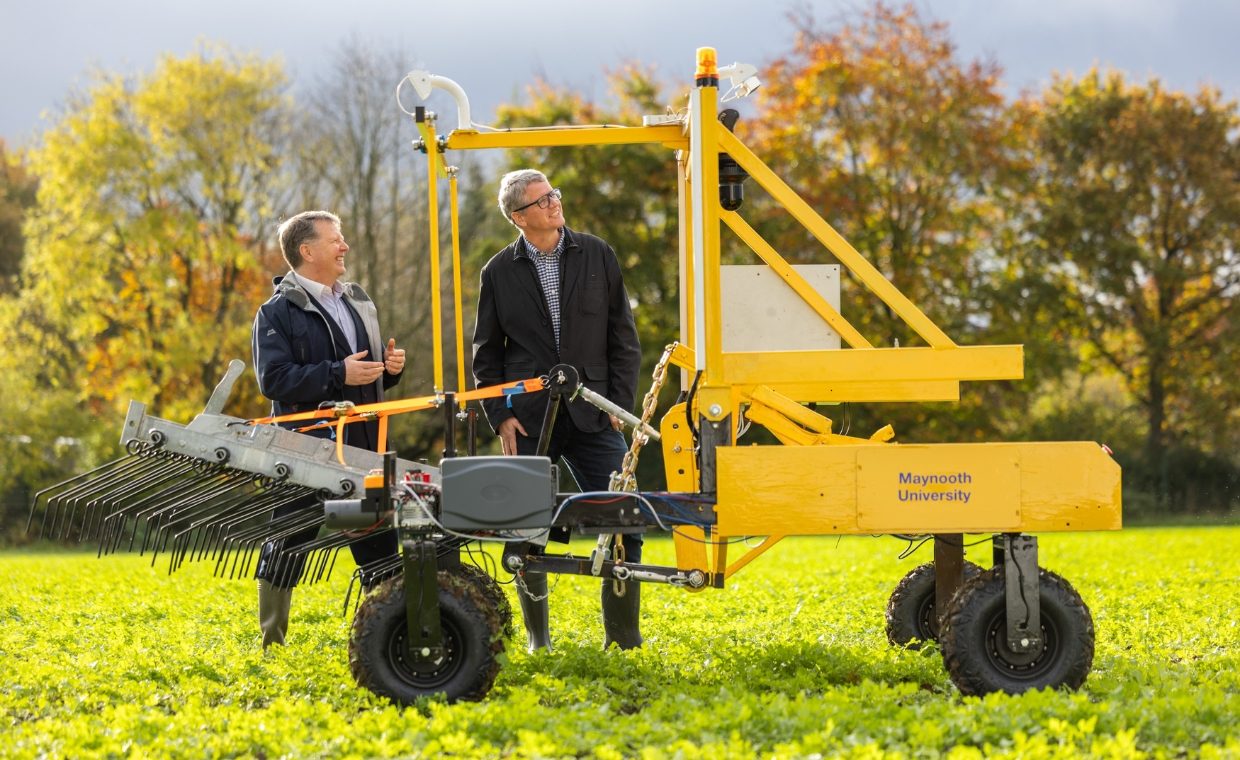 2 men standing in a field, behind a yellow seed robotic planter