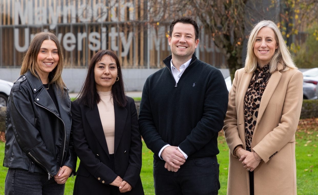 Two women dressed in black, a man in black and a woman in a camel coat in front of a building that says Maynooth University