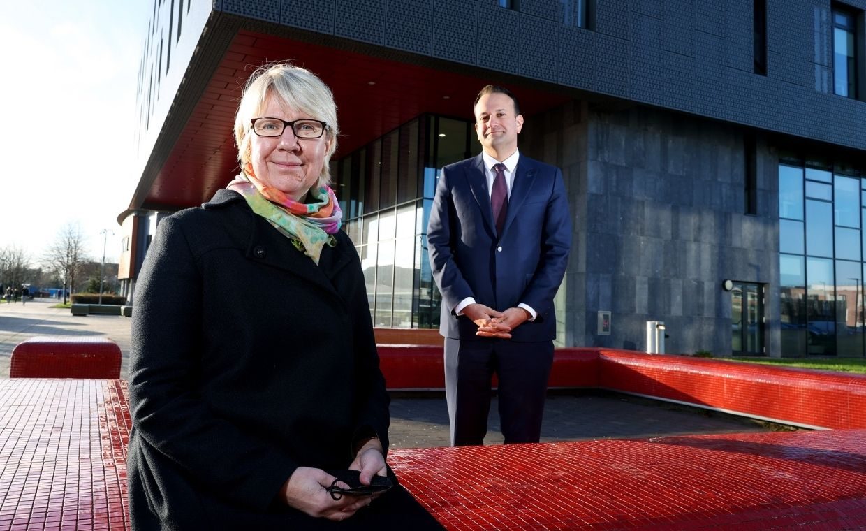 Prof Eeva Leinonen sits in the foreground on the red tiled benches outside of Eolas building, Tanaiste Leo Varadkar stands behind her.  Both are looking to camera