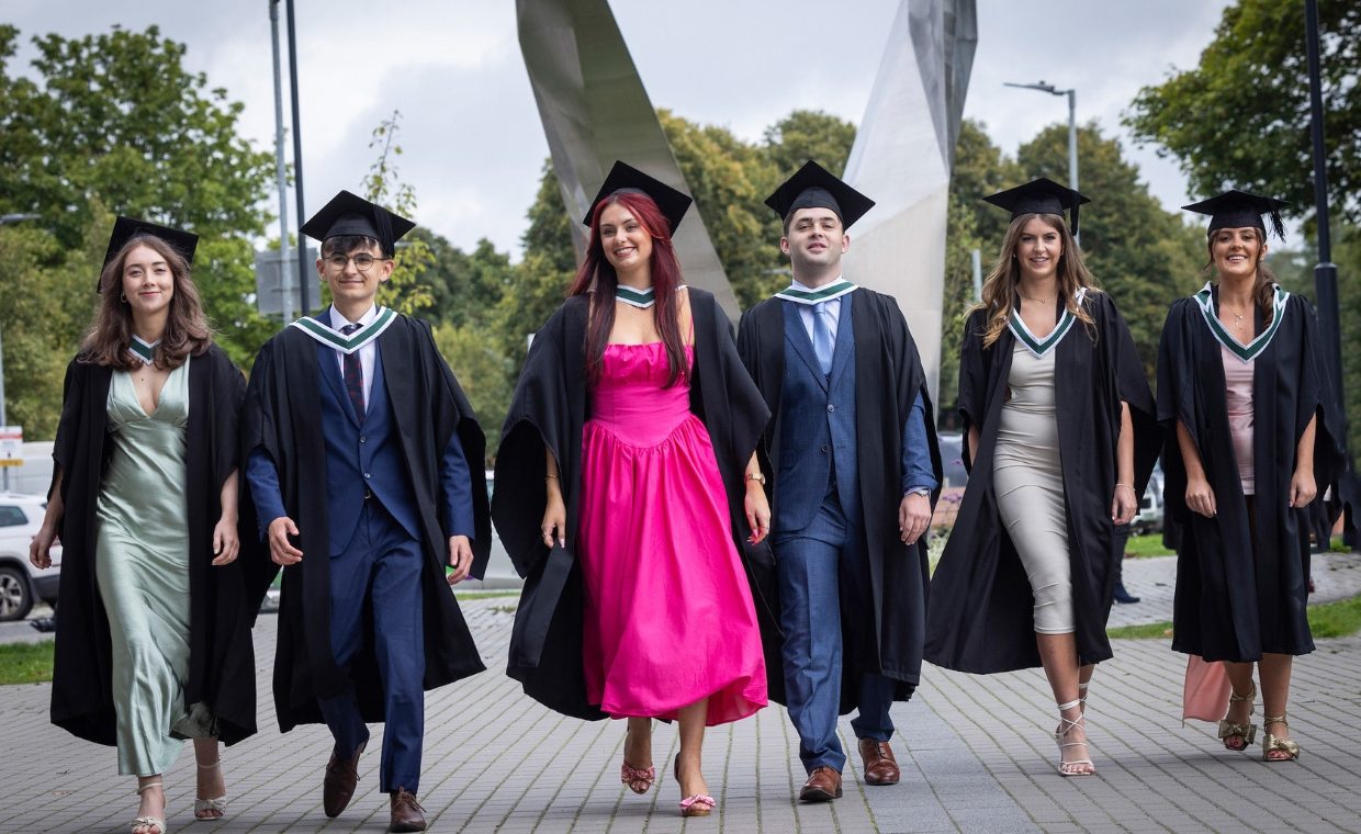 Six graduates walking in a row, girls in a pink dress in the middle