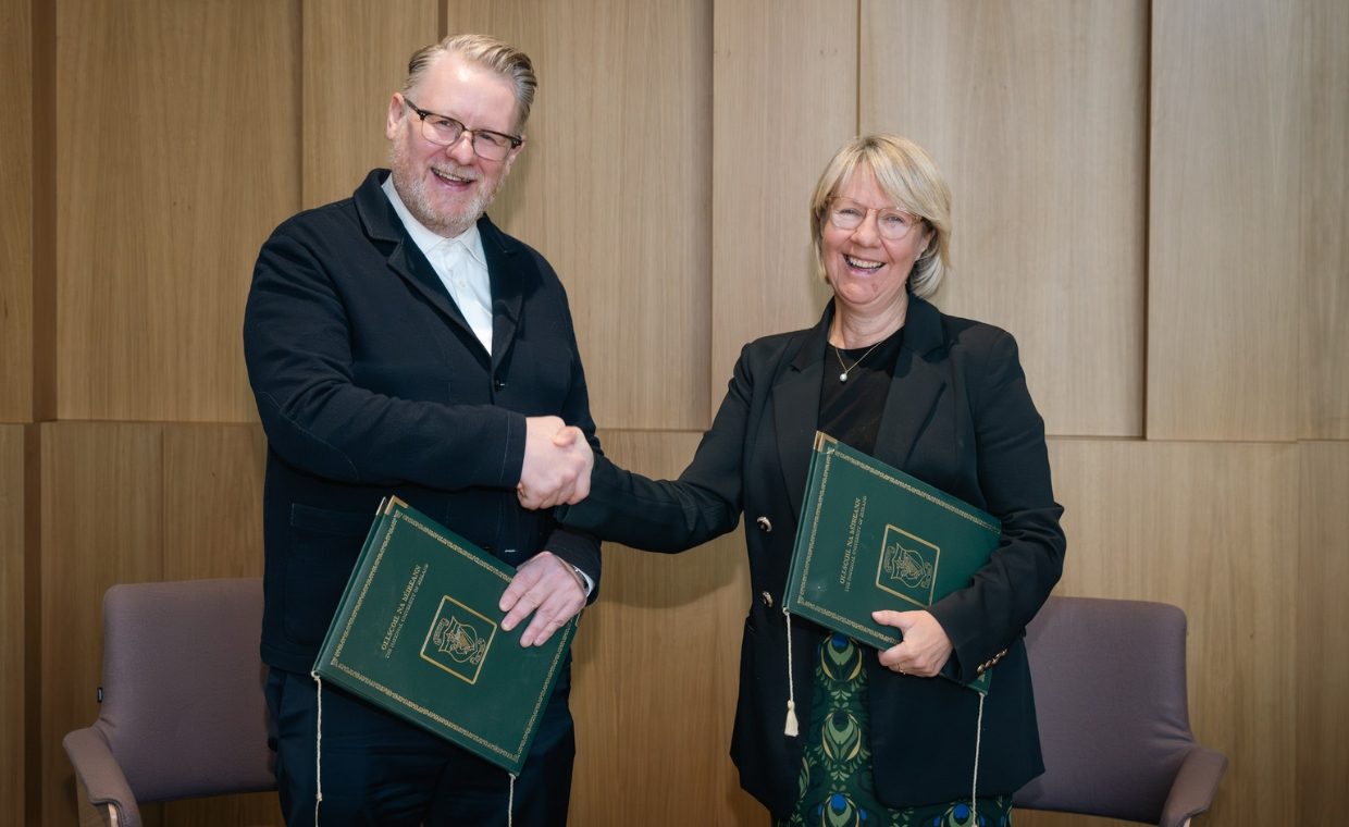 Tall man with glasses in a black jacket, holding a green folder, shakes hands with a woman in black, also holding a green folder