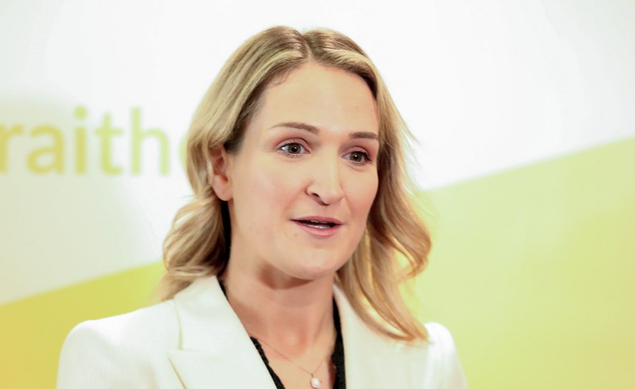 A blonde haired woman in a white blazer speaking to camera - yellow and white backdrop