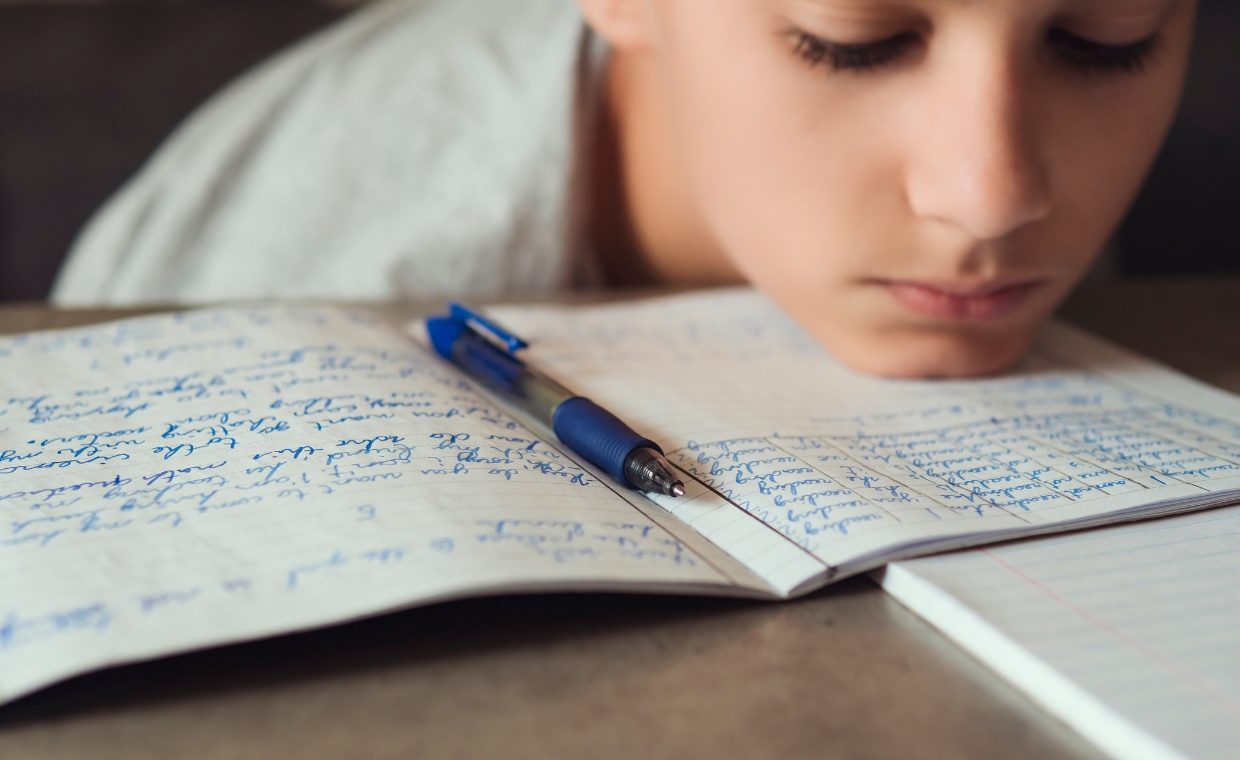 Child with sad face and head resting on table. Book with writing and a pen