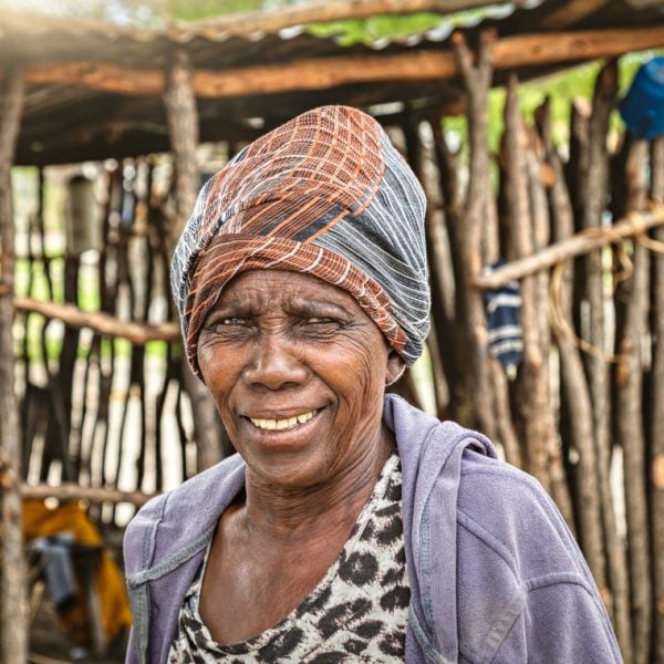 Older African woman wearing a head dress stadning in front of a hut made of sticks