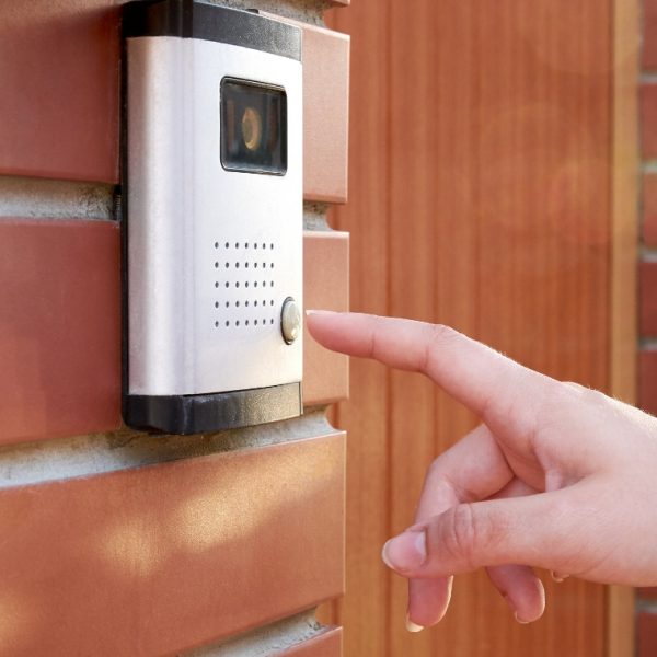 Person pushing button on white smart doorbell, red brick house, brown door