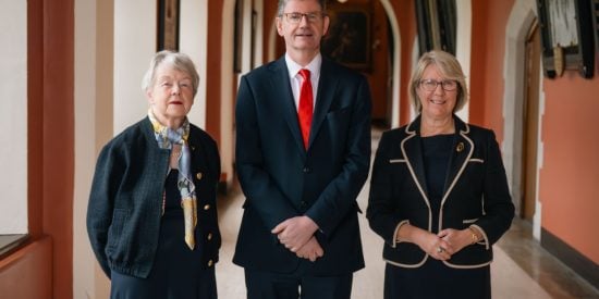 2 women and 1 man. The 2 women dressed in black stand either side of the man. Man in black suit, white shirt and red tie.