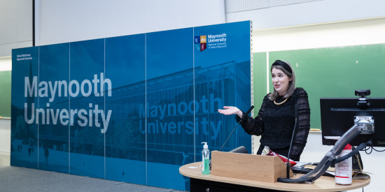 Holly Foley speaking at the STEMP Inc graduation in front of a Maynooth University backdrop