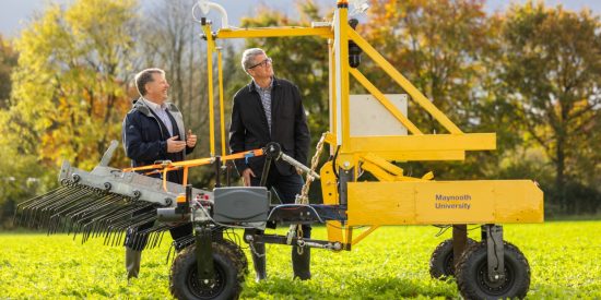 2 men standing in a field, behind a yellow seed robotic planter