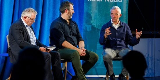 3 men on a panel discussion, blue background, left man in a navy suit, white shirt, middle man wearing navy shirt and green trousers and man to the right wearing a navy cardigan 