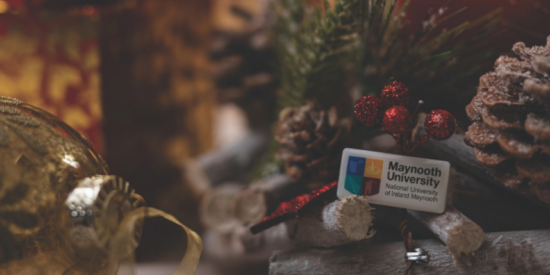 Maynooth University logo enamel pin against a background of Christmas lights and decoration