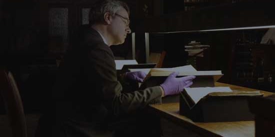 David Stifter, wearing vibrant purple gloves, sits at a bench in the Russel Library and reads a book