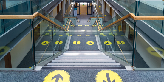 Social distancing signage on the stairs in Iontas showing which side to walk up and down. The stickers are black arrows on yellow circles