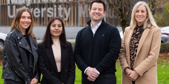 Two women dressed in black, a man in black and a woman in a camel coat in front of a building that says Maynooth University