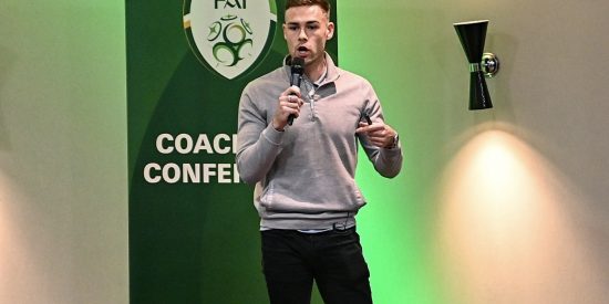 Dr Liam Sweeney, Standing with a microphone, in front of the FAI pop up stand,  speaking to an audience