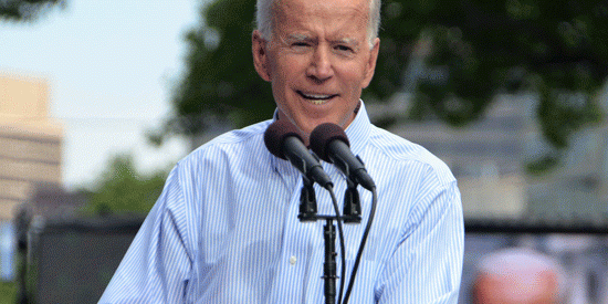Joe Biden, visible from the waist up, in a blue shirt stands at a podium and speaks into the microphone, he is smiling