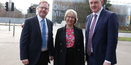 3 people posing for camera, in front of large building with font Ollscoil Mha Nuad. Man to left in dark suit, white shirt, blue tie, glasses and brown hair. Women in middle dark suit with pink, blue and green floral top, with glasses and blonde hair. Man to the right, navy blue suit with lilac shirt and purple tie. Tall man