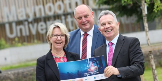 Smiling woman holding a report with two men with Maynooth University in the background