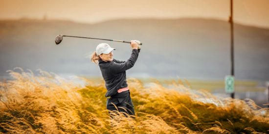 Woman golfer dressed in black with a white cap swinging out of the rough