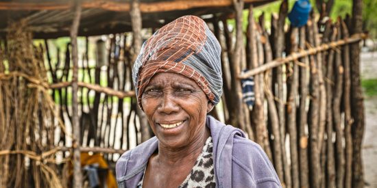 Older African woman wearing a head dress stadning in front of a hut made of sticks